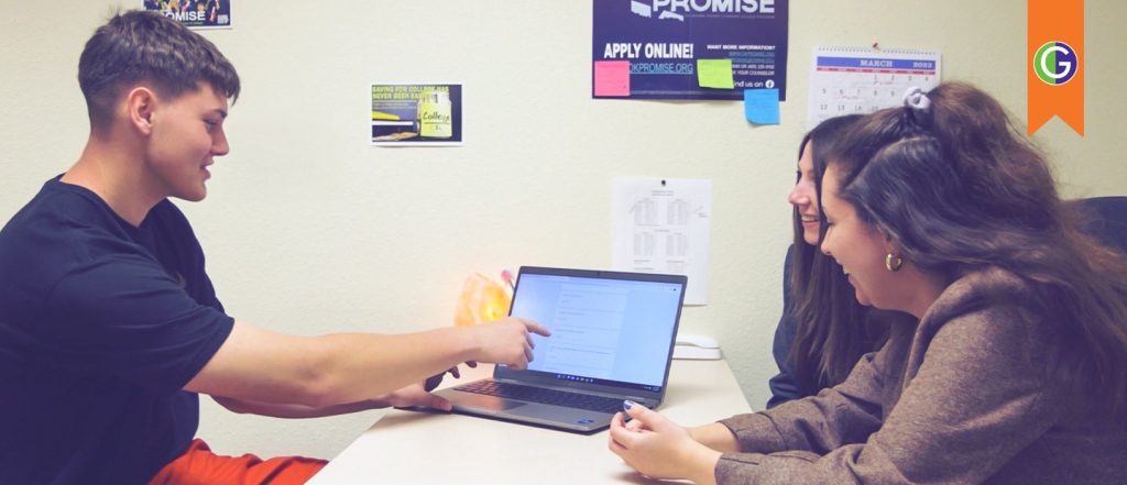 Current image: In this image, two high school students are gathered around a desk discussing financial planning with a GEAR UP Site Director while reviewing information on a laptop. At the top right, an orange ribbon with the green and purple GEAR UP logo is overlayed on the photo. The image has an Oklahoma's Promise poster on the wall behind the students that reads: "APPLY ONLINE! OKPROMISE.ORG." Another poster to the left reads: “SAVING FOR COLLEGE HAS NEVER BEEN EASIER.”