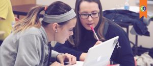 Graphic features a photo of two high school students in a classroom, seated closely and focused on a white laptop, and at the top right, an orange ribbon features the GEAR UP green and purple logo. The two female students in the photo are discussing their college application essays and preparing to start their college journey.